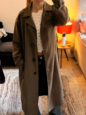 Brun/grön trenchcoat med stora knappar - Stilren brun trenchcoat med klassisk krage och stora svarta knappar framtill. Lång modell med avslappnad passform och fickor på sidorna. Sååå fin nu till våren och sommaren! Knappt använd 💕🌼🌼🌼🌼