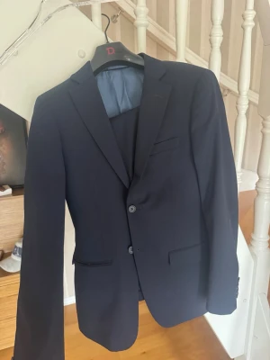 Ull dressman kostym med tvåknäppt kavaj - Snygg mörkblå kostym med klassisk tvåknäppt kavaj och matchande byxor. Kavajen har smala slag, tre fickor fram och knappar vid ärmslut. Perfekt för dig som vill ha en stilren och tidlös look.