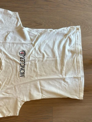 Vit Moncler t-shirt med logga - Snygg vit t-shirt från Moncler med klassisk rund hals och broderad logga på bröstet. Tillverkad i mjuk bomull som känns skön mot huden. Perfekt till jeans eller shorts för en clean och stilren look.