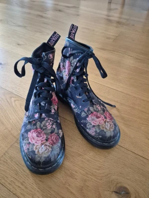 Blommiga kängor från Dr. Martens st 37 - Säljer ett par svarta kängor från Dr. Martens med färgglatt blommönster i rosa och beige toner. Klassisk snörning, robust sula. Materialet är tyg med tryckt mönster och svart gummisula. Har slitningar där trycket bleknat något, men i bra skick. 