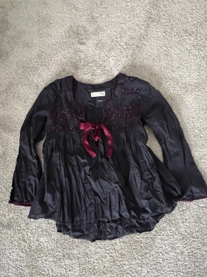 Mörklila blus med rosett från Odd Molly - Supersöt mörklila blus från Odd Molly med långärmad design och dekorativ rosett framtill. Blusen har fina broderade detaljer upptill och är gjord i ett lätt, flowy material. Perfekt för dig som gillar boho-vibbar och snygga detaljer.