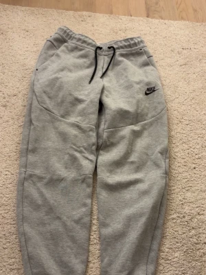 Gråa Nike teech byxor  - Snygga gråa Nike teech byxor i storleken XS. Byxorna har en svart logga på vänster ben. Byxorna har sidofickor + den klassiska dragkedje fickan. Byxan är tillverkade i mjukt sweatshirtmaterial som är perfekt för en chill dag eller ett träningspass. Klassisk och avslappnad passform.