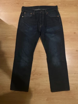 Mörkblå Levi's straight fit jeans - Säljer ett par klassiska mörkblå jeans från Levi's med raka ben och fem fickor. Jeansen har kontrastsömmar och den ikoniska läderlappen bak i midjan. Tillverkade i slitstark denim, perfekta för en avslappnad och tidlös stil. obs! Dessa är omsydda till 30 i längd och väldigt slarvigt som man såg på bilden.