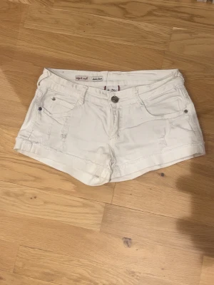 Vita denimshorts från Miss Two - Säljer ett par vita denimshorts från Miss Two med klassisk femficksdesign och knappgylf. Shortsen har en lågmidjad passform och är perfekta för varma dagar. Snygga att matcha med en enkel t-shirt eller linne.