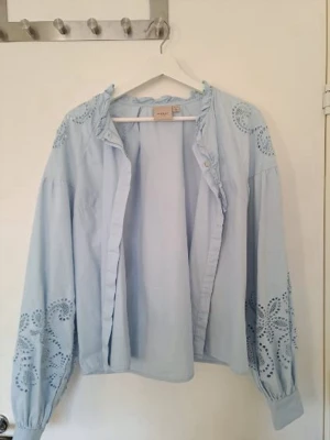 Ljusblå blus med broderade detaljer - Supersöt ljusblå blus från Vila med broderade hålmönster på ärmar och axlar. Blusen har volangkant vid kragen och knappar framtill. Använd endast 1 gång och säljer nu pga att den tyvärr va för stor.