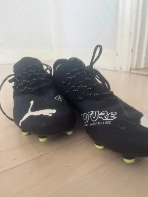 Puma Future Z Svarta Fotbollsskor - Säljer ett par svarta soprena Puma Future Z fotbollsskor med vita detaljer och logga. Skorna har FG-dobbar i neonfärg och en strumpliknande passform för extra komfort och kontroll på planen. Perfekta för dig som vill ha stil och prestanda på matchen.
