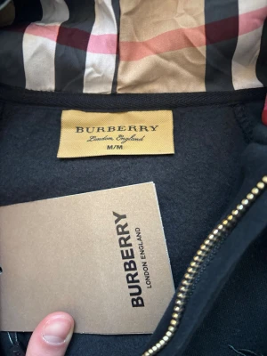 Svart hoodie med dragkedja från Burberry - Snygg svart hoodie från Burberry med dragkedja och klassiskt rutigt foder i huvan. Broderad Burberry-logga på bröstet och guldfärgade detaljer på snörena. Perfekt för dig som vill ha en exklusiv och stilren look.