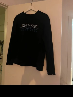 Svart sweatshirt från Hugo Boss - Snygg svart sweatshirt från Hugo Boss med stort BOSS-tryck på bröstet och texten 'Hugo Boss' under. Tröjan har rund halsringning och är i slim fit-modell. Perfekt för en clean och stilren look.