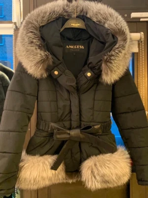 Svart pälsjacka från AmoreSS - Snygg svart pälsjacka från AmoreSS med stora fluffiga fuskpälsdetaljer i grått på huva och nederkant. Jackan har bälte i midjan för en markerad siluett och quiltad design. Perfekt för kalla vinterdagar och ger en lyxig känsla.