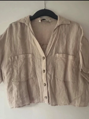 Ny. Beige linneskjorta med knappar - Snygg beige skjorta i linne med kort ärm och två stora bröstfickor. Skjortan har en avslappnad passform, klassisk krage och knäppning framtill. Perfekt för en chill och stilren look. Oversize, kan även passa S och M.