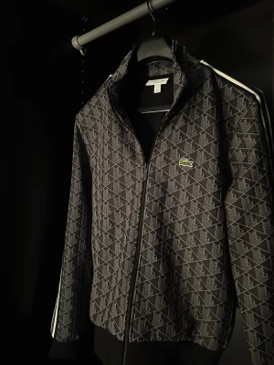 Svart track jacket från Lacoste - Säljer en svart zip-tröja från Lacoste med diskret geometriskt mönster och vita ränder längs ärmarna. Tröjan har hög krage, ribbade muddar och det klassiska Lacoste-logotypen på bröstet. Perfekt för dig som gillar sportig och stilren design. Haft på mig den en gång , och den har blivit mindre på mig på grund av gymmet 🥲 , Så nu är det dags och sälja den🫡. 100% äkta ✅ Pris kan diskuteras