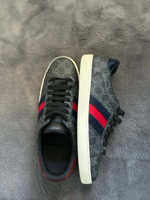 Gucci sneakers med GG-mönster - Snygga Gucci sneakers i mörkgrått med klassiskt GG-mönster, blå och röda ränder på sidan samt metallicröd och metallicblå detalj på hälen. Skorna har vita sulor och svarta skosnören. Materialet är en mix av canvas och läder.