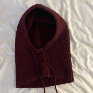 Vinröd stickad balaclava med snören - Mysig vinröd stickad balaclava i ribbat material. Huva med dragsko framtill som ger en cool och avslappnad look. Ingen stolek. Köpte för 450