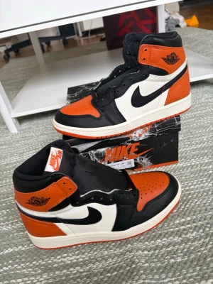 Nike Air Jordan 1 Shattered Backboard - Grymma Nike Air Jordan 1 Shattered Backboard med svart, orange och vit färgkombination. Klassisk high-top siluett med läderpaneler, svart swoosh och orange detaljer på tå, häl och yttersula. Perfekta för dig som vill sticka ut med en ikonisk sneaker.