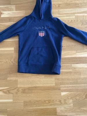 Blå hoodie från GANT med logga - Snygg blå hoodie från GANT med broderad logga och amerikanskinspirerat sköldmärke på bröstet. Klassisk känguruficka framtill och huva. Tillverkad i mjukt bomullsmaterial som är skönt att ha på sig. Perfekt för en avslappnad och sportig stil.