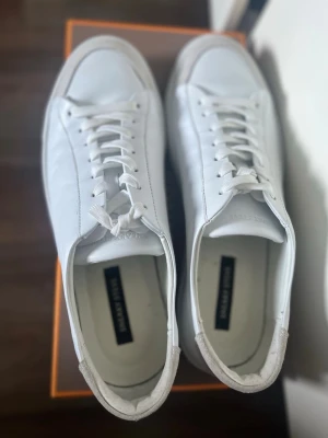 Vita sneakers från sneaky steeve - Stilrena vita sneakers från sneaky steeve  i slätt läder med klassisk låg profil och vita snören. Skorna har en tjock vit gummisula och diskret branding på plösen. Perfekta för en clean och modern look.