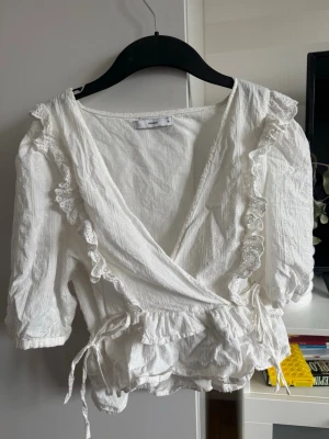 Vit omlottblus med volanger och spets - Söt vit omlottblus med volangdetaljer och knytband i midjan. Blusen har djup V-ringning, spetskant och trekvartsärm. Perfekt att styla med jeans eller kjol. Använt någon gång men har legat i garderoben ett tag, för liten i bysten för mig