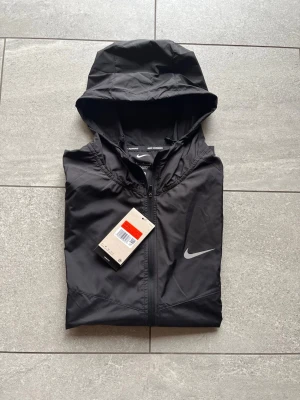 Svart Nike runner vindjacka - Snygg svart vindjacka från Nike med huva och dragkedja framtill. Storlek S. Inga defekter alls och lappen följs med.