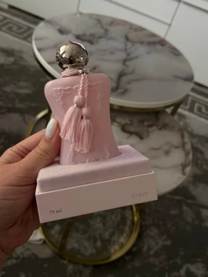 Parfym Parfums de Marly Delina 65 ml - Lyxig parfymflaska i ljusrosa från Parfums de Marly, modellen Delina. Flaskan har en elegant form med dekorativa tofsar och silverfärgat lock. Volymen är 75 ml och designen är feminin och exklusiv, perfekt som statement på sminkbordet.