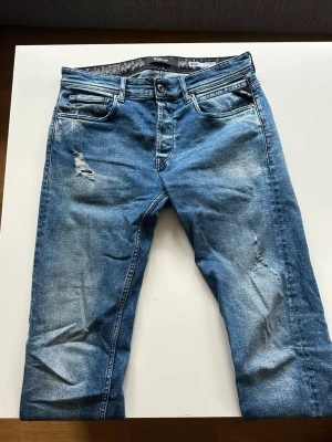 Blå slitna jeans från Replay - Säljer ett par blå jeans från Replay med slitningar och distressed detaljer på låret och knät. Jeansen har en snygg tvättad look och raka ben. Perfekta för en avslappnad stil. Modell Grover straight fit, W31 L34, Sitter som W30