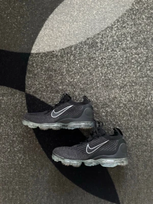 Nike Vapormax  - Svarta Nike Air Vapormax sneakers med stickad ovandel och tydlig vit swoosh på sidan. Skorna har genomskinlig bubbelsula och svarta snören. Ovandelen är i mesh för extra andningsförmåga och komfort. Perfekta för en sportig och modern look. Storlek 44 1/2. Helt Nya och oanvända