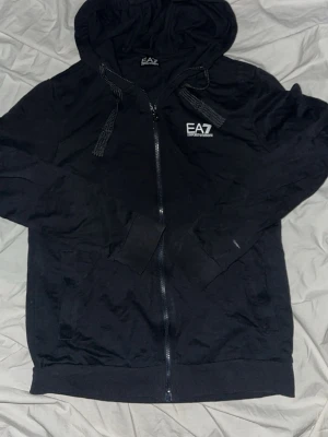 Svart EA7 Emporio Armani hoodie - Svart hoodie med dragkedja från EA7 Emporio Armani. Har huva med snören, vita logotypdetaljer på bröstet och reflexränder på ryggen. Perfekt för en sportig och stilren look. Tillverkad i mjukt material för extra komfort.