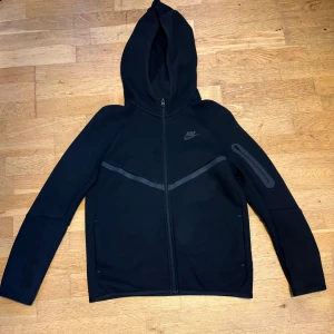 Svart Nike hoodie med dragkedja - Svart hoodie från Nike med hel dragkedja framtill och huva. Tröjan har en diskret Nike-logga på bröstet, en snygg diagonal detalj över bröstet och en ficka med dragkedja på ena ärmen. Perfekt för en sportig och avslappnad stil.
