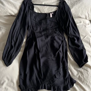 Svart omlottklänning med volang - Supersnygg svart kortklänning från Nelly med långa puffärmar, omlottdetalj och volangkant nertill. Klänningen har rynk vid bysten och dragkedja i ryggen. Perfekt för dig som gillar en feminin och trendig look.