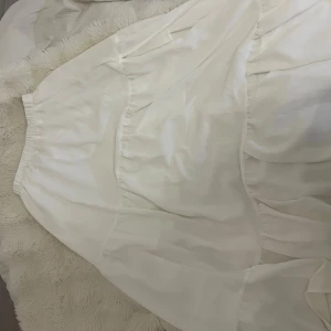 Vit långkjol med volang - Säljer en vit långkjol med volangdetaljer och resår i midjan. Kjolen har en luftig och flowy känsla, perfekt för varma dagar. Materialet känns lätt och svalt mot huden.