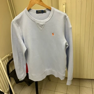 Ljusblå sweatshirt från Polo Ralph Lauren - Säljer en stilren ljusblå sweatshirt från Polo Ralph Lauren med klassisk rund halsringning och broderad logga i orange på bröstet. Tröjan har långa ärmar och är tillverkad i mjuk bomull, perfekt för en chill och clean look.