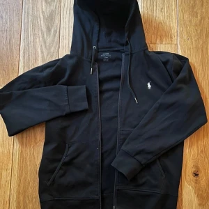 Svart hoodie från Polo Ralph Lauren - Svart hoodie från Polo Ralph Lauren med dragkedja, huva med snörning och klassisk vit logga på bröstet. Mjuk bomullskvalitet och två fickor framtill. Perfekt för en chill och stilren look.