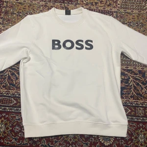 Vit sweatshirt från BOSS i storlek L - Vit sweatshirt från BOSS med svart logotyptryck på bröstet. Klassisk rund halsringning och långa ärmar. Tröjan är i mjuk bomull och har ribbade muddar vid ärmslut och nederkant. Perfekt för en clean och stilren look.
