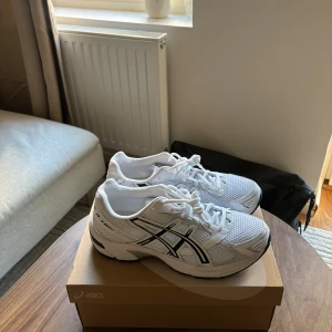 Vita Asics GEL-1130 sneakers strl 42 - Säljer ett par fräscha vita Asics GEL-1130 sneakers i storlek 42. Skorna har mesh- och syntetpaneler, svarta detaljer och klassisk Asics-logga på sidorna. Perfekta för dig som gillar sportig stil och vill ha bekväma skor med snygg design.