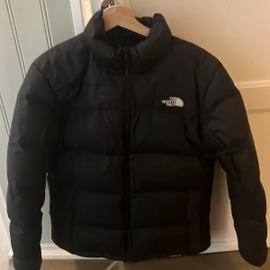 Svart dunjacka The North Face 700 - Svart dunjacka från The North Face, modell 700. Jackan har en klassisk pufferstil med hög krage, broderad logga på bröstet och ärmarna samt dragkedja framtill. Perfekt för kalla dagar och riktigt snygg till streetwear.