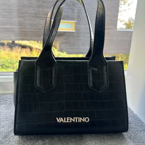 Svart handväska från Valentino - Snygg svart handväska från Valentino med croco-mönstrad yta och guldfärgade detaljer. Insidan har fack med dragkedja för småsaker. Perfekt för dig som vill ha en stilren och trendig handväska. Endast använd vid enstaka tillfällen