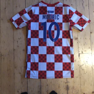 Kroatien Modric 10 fotbollströja - Säljer en klassisk Kroatien fotbollströja med Modric och nummer 10 på ryggen. Tröjan har rödvitt rutigt mönster, blå siffror och Kroatiens landslagsemblem på bröstet. V-ringad hals och korta ärmar. Perfekt för dig som vill stötta laget med stil.