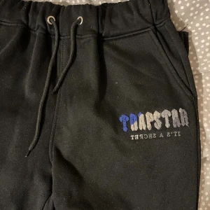 Svarta mjukisbyxor från Trapstar - Svarta mjukisbyxor från Trapstar med broderad logga i blått, grått och vitt på ena benet. Byxorna har dragsko i midjan och är tillverkade i mjukt material som känns skönt mot huden. Perfekta för chill eller streetwear.