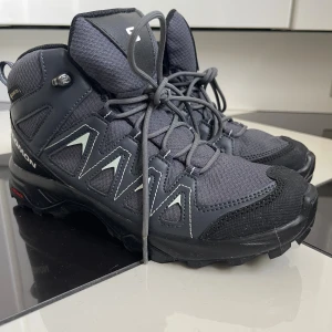 Salomon X Braze GTX kängor svarta - Salomon X Braze GTX kängor i svart och mörkgrått med robust snörning och grov sula. Skorna är tillverkade i syntet och skinn, har vattentätt Gore-Tex-material och detaljer i vitt. Perfekta för utomhusaktiviteter och äventyr.