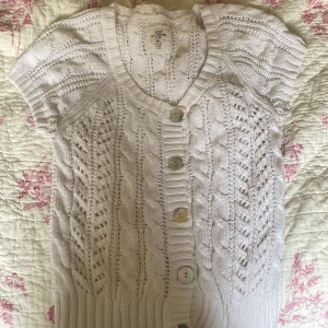 Vit stickad kofta från L.O.G.G. - Kortärmad vit stickad kofta från L.O.G.G. med snyggt kabelstickat mönster och stora pärlemorknappar framtill. Koftan har ribbade kanter och en mjuk, luftig känsla som passar perfekt till lager-på-lager-stil.