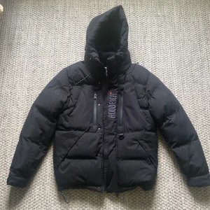 Svart pufferjacka från Hoodrich storlek S-M - Svart pufferjacka från Hoodrich med huva och tryck på både bröst och rygg. Använd en gång helt nyskick riktigt varm ock skön, storlek S men passar även M. 