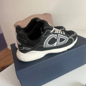 Svarta Dior sneakers med vita detaljer - Säljer ett par svarta Dior sneakers med vita och grå detaljer samt stora CD-loggor på sidorna. Skorna har vit sula, vita skosnören och är tillverkade i en mix av mesh och syntetmaterial. Kommer med originalkartong och extra skosnören.