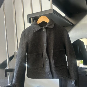 Svart ulljacka med stora fickor - Säljer en svart, kort ulljacka med klassisk krage och stora knappar framtill. Jackan har två rejäla fickor och en rak, boxig passform som ger en cool och stilren vibe. Perfekt för dig som gillar minimalistisk stil och vill ha något som funkar till många outfits.