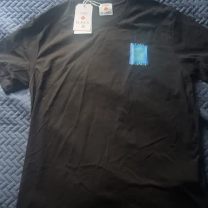 Svart Casa Blanca t-shirt med blå print - Svart t-shirt från Casa Blanca med stor blå grafisk ruta och logga på ryggen samt mindre tryck på bröstet. Klassisk passform och rund hals. Perfekt för dig som gillar stilrena men ändå unika plagg. Materialet är mjuk bomull.