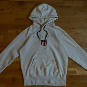 Vit hoodie från GANT med broderad logo - Vit hoodie från GANT med broderad GANT-text och sköld på bröstet. Tröjan har huva med svarta snören och en stor magficka. Tillverkad i mjuk bomull, perfekt för chill dagar. Klassisk och stilren design med snygga detaljer.