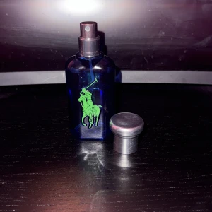 Ralph Lauren blå parfymflaska - Stilren parfym från Ralph Lauren i blå flaska med grön logga. Perfekt för dig som vill ha en ikonisk accessoar på hyllan. Passar dig som gillar klassisk och sportig stil. Fräsch doft med noter av bland annat citrus. Ungefär 40ml kvar.
