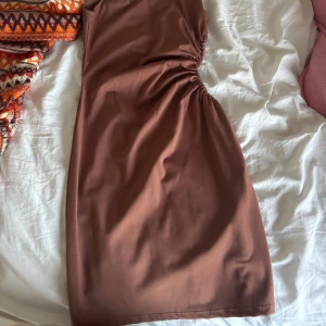 Brun kort klänning med snörning - Snygg brun kort klänning med smala axelband och cool snörningsdetalj på sidan som ger en unik form. Klänningen har en enkel, stilren look och är figurnära med lätt stretchigt material. Perfekt för dig som gillar minimalistisk stil med en twist.