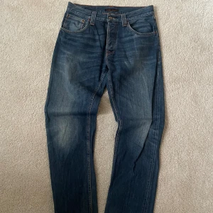 Blå jeans från Nudie Jeansco W32 L34 - Säljer ett par klassiska blå jeans från Nudie Jeansco med snygga slitningar och kontrastsömmar. Modellen har raka ben, fem fickor och coola broderade bakfickor. Perfekt passform och tidlös stil för dig som gillar jeans med attityd.