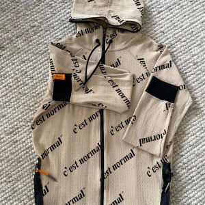 Beige hoodiejacka från C'est Normal - Beige hoodiejacka från C'est Normal med svart textprint över hela jackan. Jackan har dragkedja, huva med snörning och svarta detaljer vid ärmsluten. Tillverkad i bomull och polyester med en skön, lite strukturerad känsla.