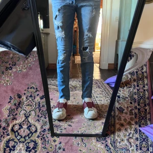 Ljusblå slim jeans med slitningar 32/30 - Säljer ett par ljusblå skinny jeans med rejält slitna detaljer och hål på båda benen. Jeansen har klassisk femficksdesign, dragkedjegylf och är tillverkade i stretchigt denimtyg för en tight passform. Perfekta för dig som gillar en edgy och trendig look. Modellen är 178cm 70kg midja 37 längd 97cm (15)