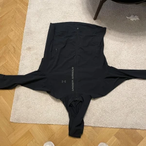 Svart vindjacka från Under Armour - Svart vindjacka från Under Armour med dragkedja framtill och logga längs bröstet. Jackan har lång ärm, hög krage och två sidofickor med dragkedja. Perfekt för aktiva dagar och sportiga outfits.